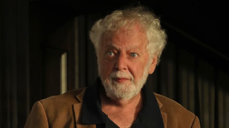 An older man with white curly hair and a beard wears a black collared shirt under a brown blazer.