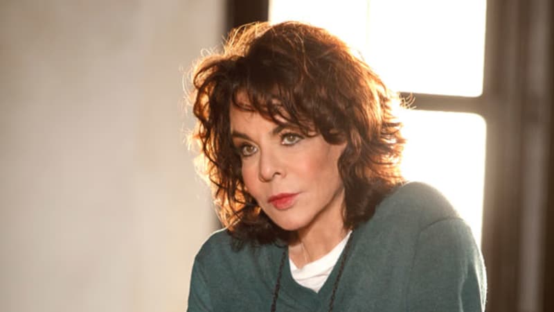 An older woman with light skin and touseled, wavy brown hair wears a dark green sweater and has her glasses hanging around her neck on a string. She leans against a kitchen chair.