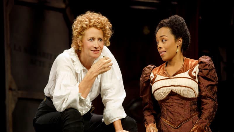 A woman with short curly blond hair wearing a white Shakespearean blouse and black trousers straddles a marble bench and speeks to a woman in a fancy Shakespearean dress who kneels next to the bench.