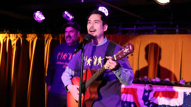 A man plays a guitar behind a microphone at an event. Behind him is a yellow curtain and an American bunting flag. Another man in a matching graphic tee stands to the side.