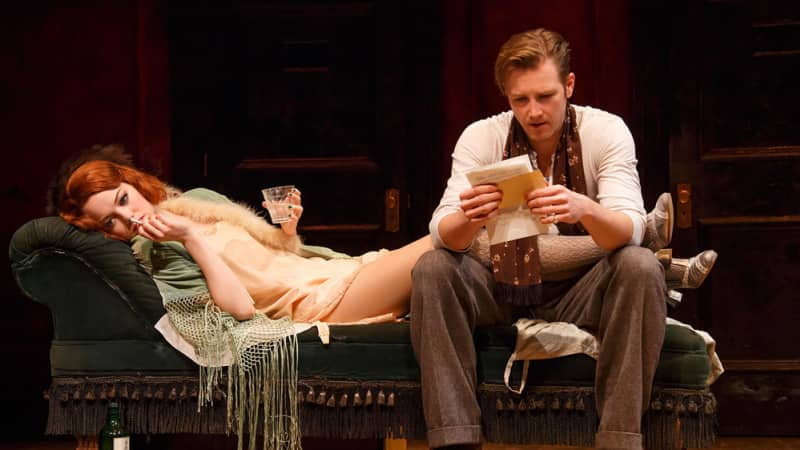 A woman with short red wavy hair lays on a chaise lounge with a cigarette and drink in her hands. A man sits at the end and reads a letter.