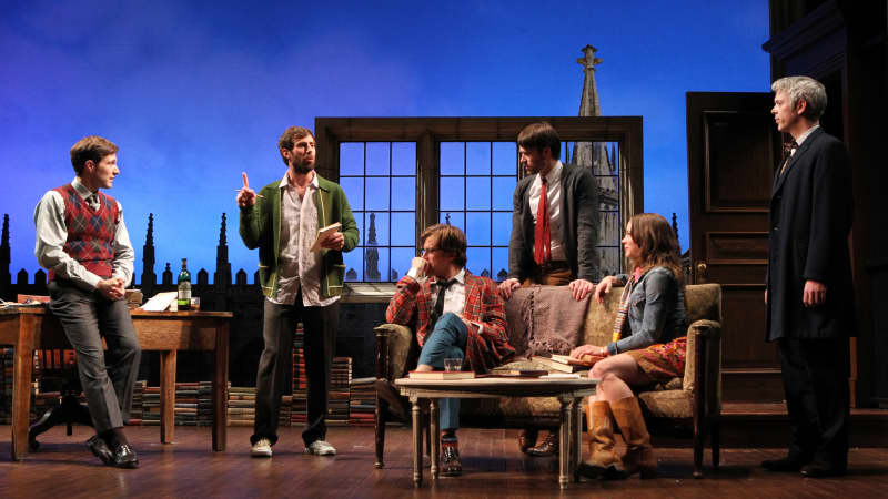 A group of men and one woman in a loft all listening to one man with a notebook speak. 