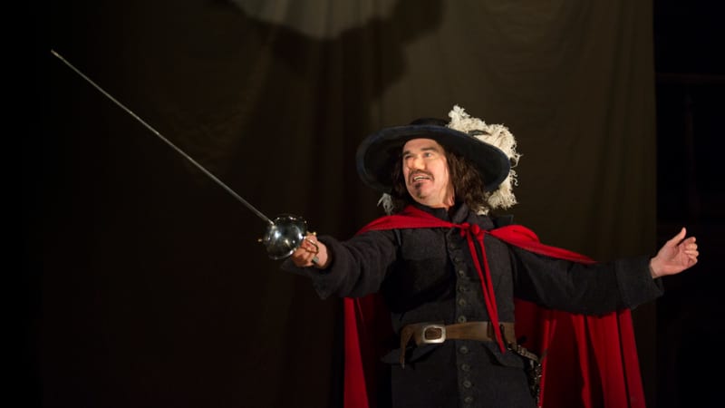 A man with long brown hair and a very large nose gestures his hands outwards with his sword. He wears a feathered cap, black 17th century clothes, and a red cape.