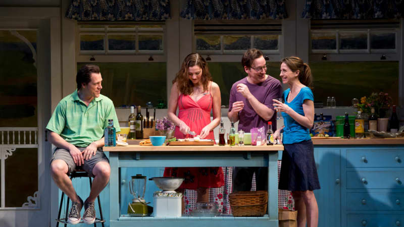Four actors on a blue kitchen set with checkered flooring. A man sits on a stool next to an island where a woman prepares food. A man and woman hold drinks and stand near the island.