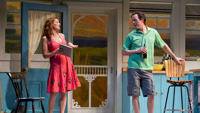 A woman in a bright pink dress talks to a man in a green polo shirt holding a beer bottle in the middle of a blue kitchen.