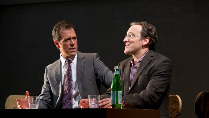 Two men in business suits sit at a table talking with drinks.