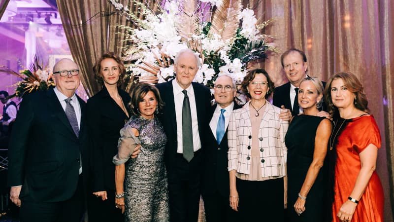 A group of people in formal attire pose together and smile in front of a large white floral arrangement.