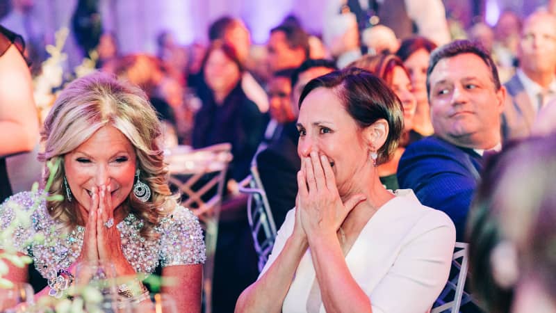 Two women in formal attire sit in a crowded ballroom and cover their mouths in amusement.