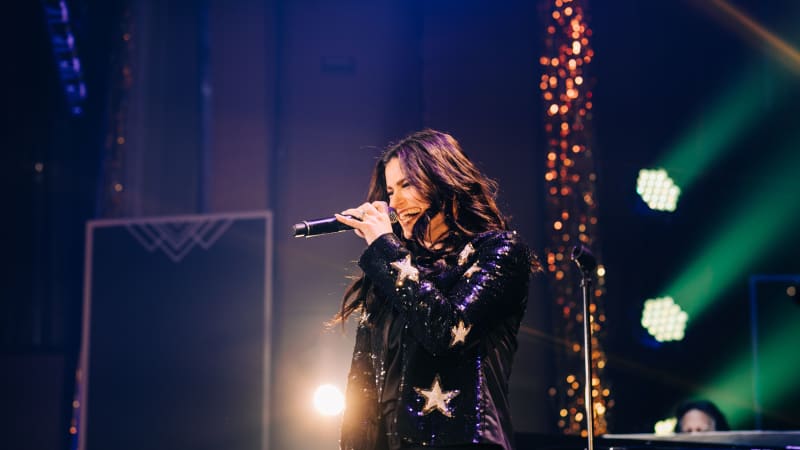 Idina Menzel sings on stage, her hair flowing in motion. She wears a sparkly blazer with stars on it.