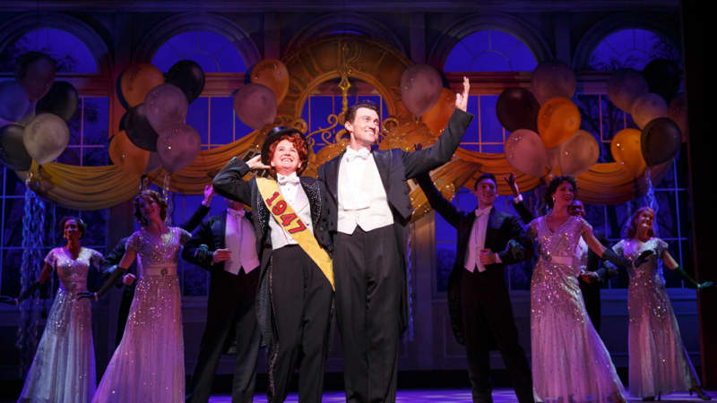 A woman and man, both in tuxedos with white bowties, stand centerstage at a celebration, surrounded by party guests in fancy outfits. The woman wears a top hat and a sash that says 1947.