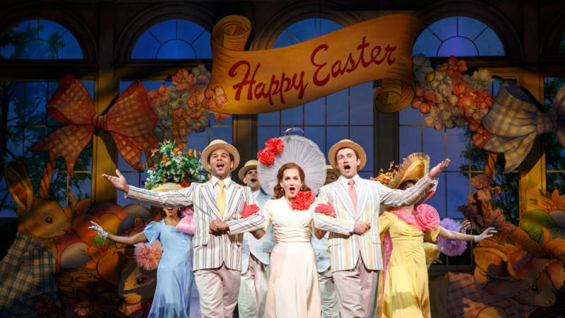 An ensemble of actors is pastel dinner suits, hats, and dresses dance in a clump with arms outstretched under a backdrop that says, Happy Easter.