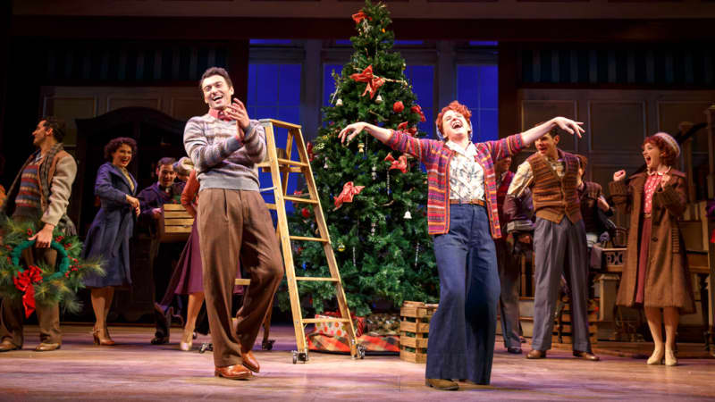 An ensemble of happy actors in 1940s clothing decorate the inn for Christmas, a giant tree in the center. A woman holds her arms outstretched at the front of the stage and a man laughs with her.