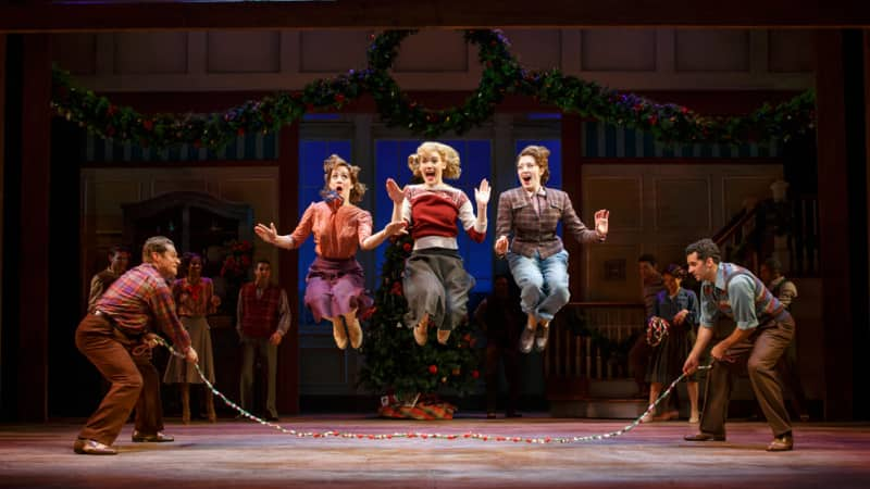 Three women in 1940s clothing jump over a Christmas-y jump rope with two men holding either side of the rope.