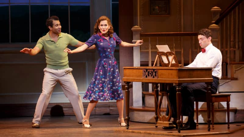 A man with a red bowtie plays at a wooden piano while a man in a green shirt and a woman in a purple dress dance next to it.