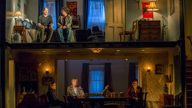 A split level set of a house. On the top in a bedroom, an older and younger man sit on the edge of a bed. Downstairs in the dining room, three people quietly sit at the table.