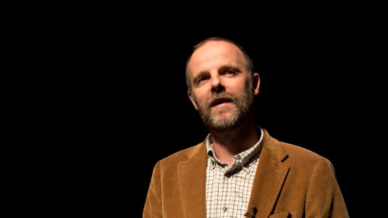 A man stands with his hand out in a spotlight wearing a corduroy tan jacket. 