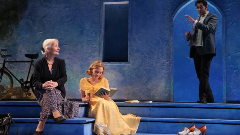 An old woman and a young blonde woman in a yellow dress reading a book sit on blue steps. Next to them, an Indian man in a casual suit talks to them.