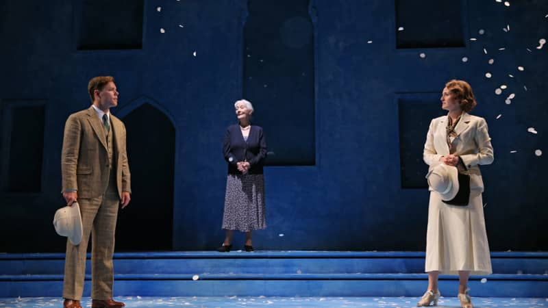 A man in a tan suit looks across the stage at a woman in a white suit dress. Between them up a few steps is an old woman. White petals fall down around them.