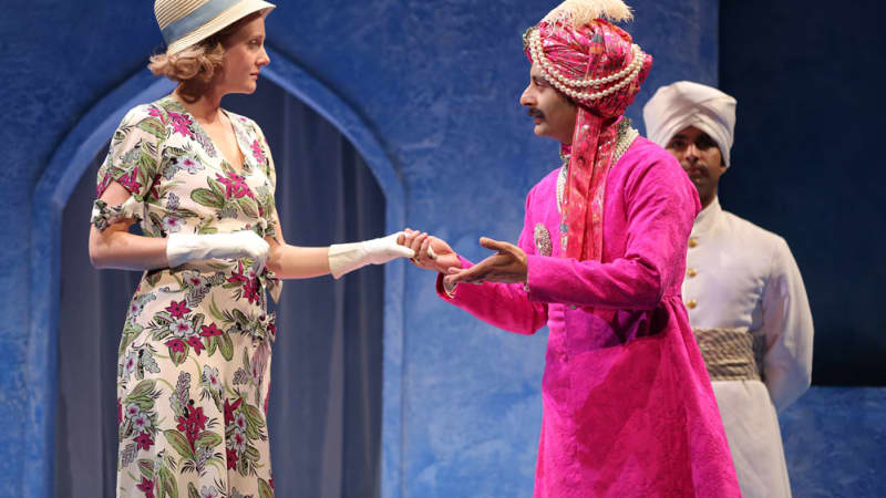 A man dressed in fancy, pink, Indian clothes and a turbin takes the gloved hand of a white woman in a floral dress.