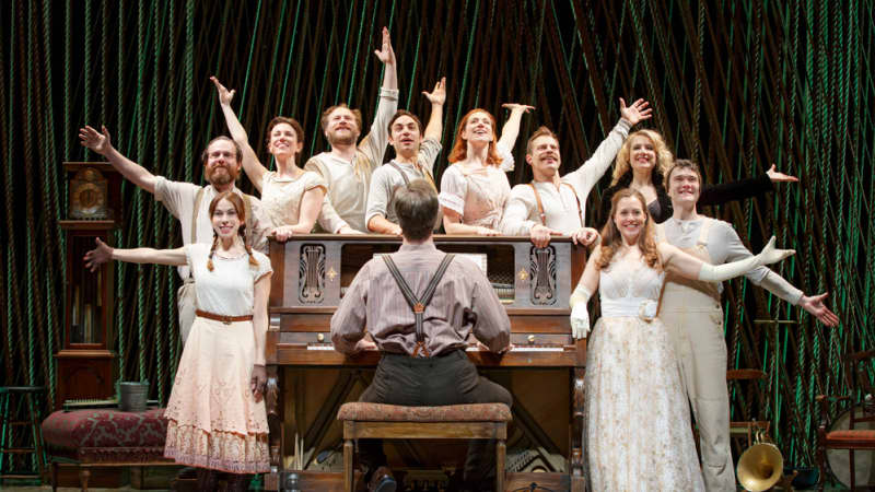 The cast, all dressed in white, pose with their arms out and stand around a man playing the piano.