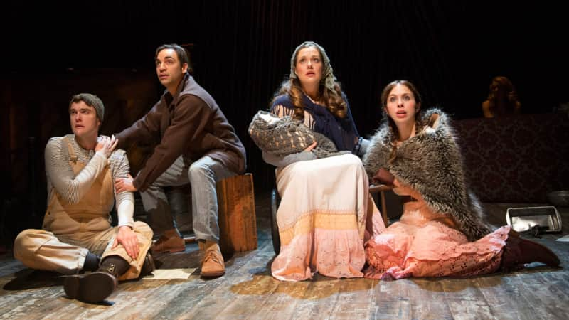 A man in overalls, a man in a brown jacket, a woman holding a baby, and a woman in a furry cape all sit together and look sad and frightened.