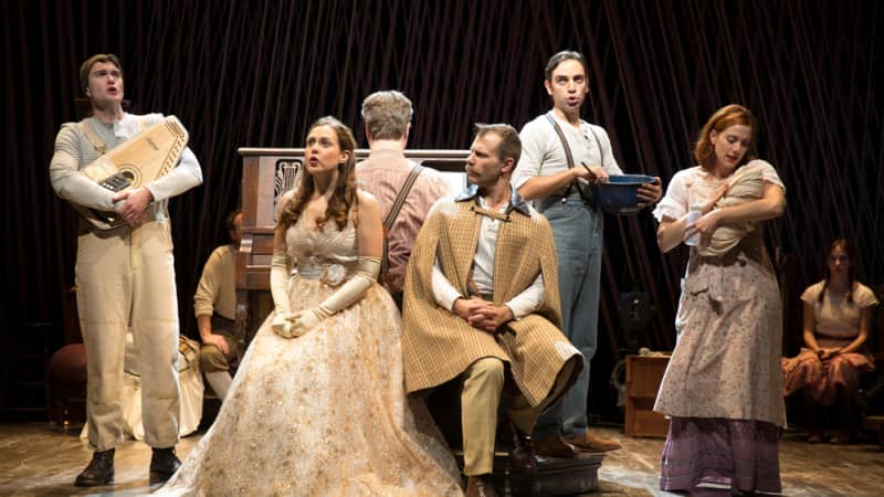 Around a man playing a piano is a young man holding a lap harp, a woman in a golden dress, a man in a cape, a man mixing a bowl, and a woman holding a baby.