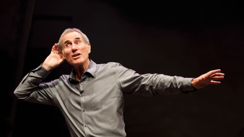 A man with gray hair wearing a gray collared shirt cups a hand around his ear dramatically, like he's listening to something off in the distance.