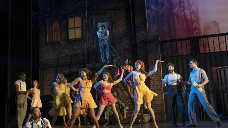 The company of KISS ME KATE. A group of women in yellow and pink dresses dance while men in t shirts and suspenders watch.