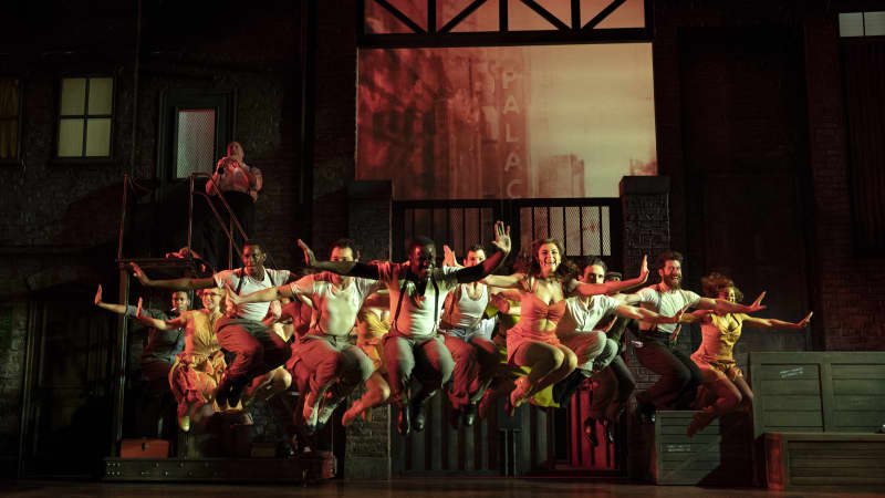 A group of dancers under red lighting jump up with their kneees tucked and arms outstretched in front of a New York City background.