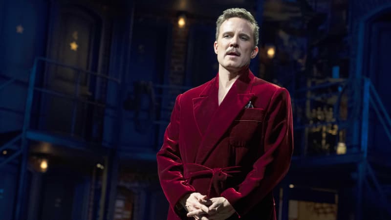 A man in a velvet red dressing gown clasps his hands together and sings on a dimly lit stage.