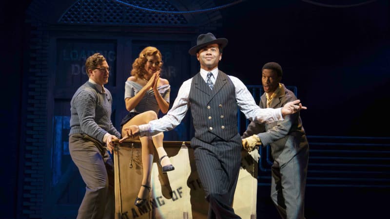 A man in a pinstriped suit and a fedora dances in front of a laundry cart with a red-headed woman sitting on top of it as two people push her across the stage.