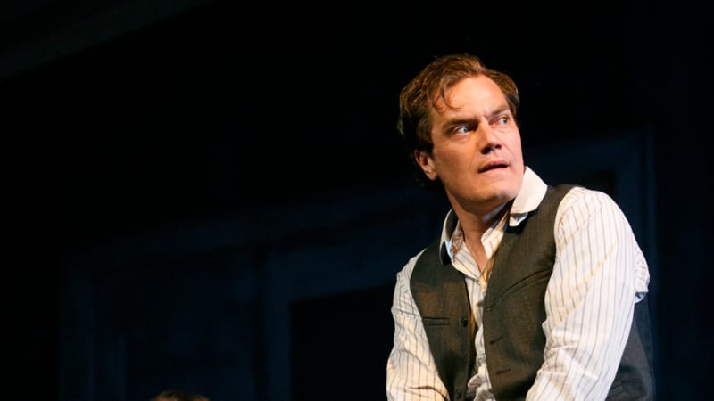 A man in a vest and collared shirt with a concerned expression leans against a wooden table. Behind him, another man in a vest sits in a low chair.