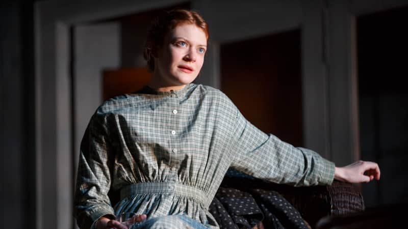 A woman in a plain, plaid, 1910s dress leans on the arm of a chair. She wears her red hair in a low bun.