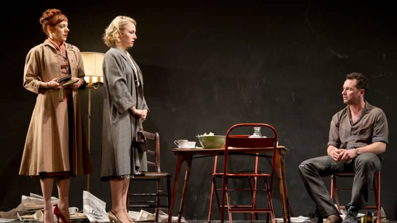 Two women in coast stand together in a dirty room over a man who seems sad sits in a chair and looks at them. 