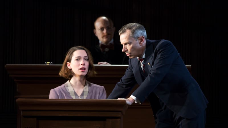 A woman with short brown hair in a purple 1930s dress sits behind a witness stand. A man in a suit leans onto the stand in an intimidating manner.