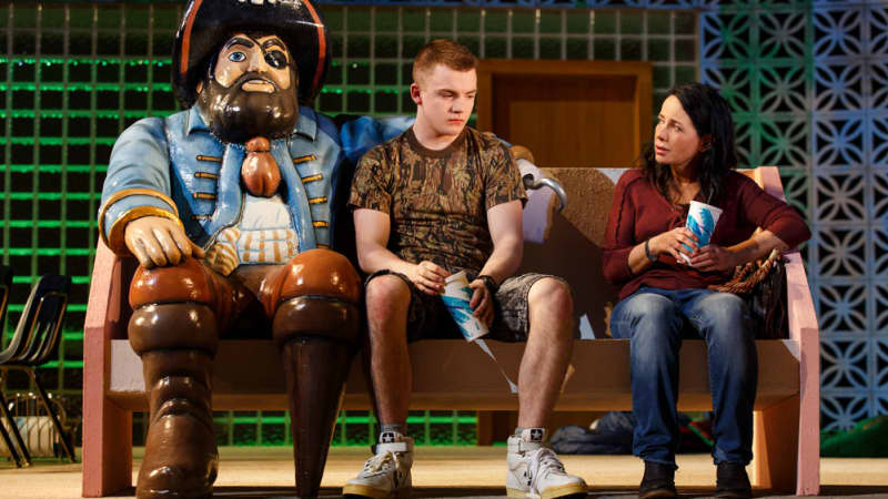 A sad young man and woman holding soda cups sit on a bench with a statue of a pirate on one end, who looks like his arm is around the young man.