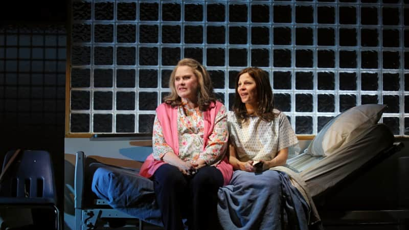 Two women sit together on the edge of a hospital bed at night. One wears a floral shirt and pink vest. The other wears a hospital gown and is under blankets.