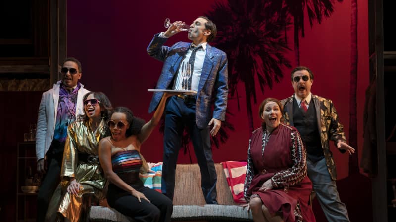 A man in a fancy suit stands on a sofa and drinks from a champagne glass, surrounded by people in sunglasses in front of a sunset palm tree background.