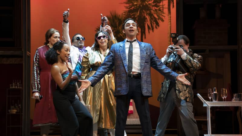 A man in a fancy suit sings center stage while fans and tourists surround him with excitement. The scene takes place in front of a sunset palmtree background.