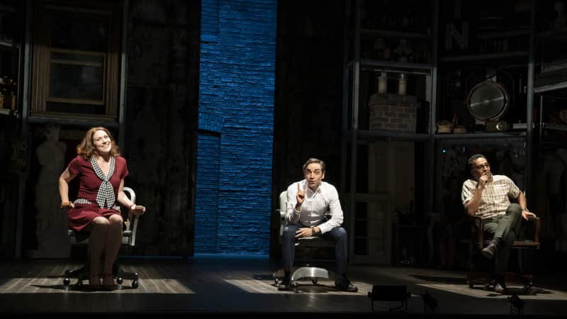 A woman and a man each sit in rolling office chairs and face the audience. They are each lit by a rectangular spotlight.