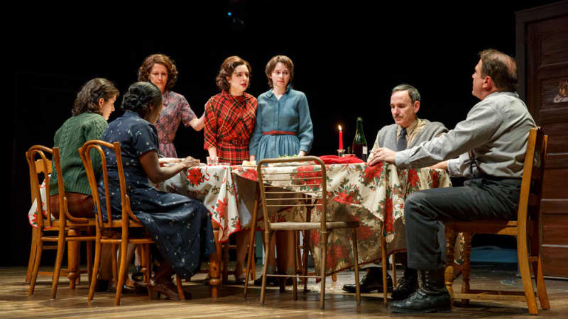 A family sits at a long dining table with multiple tablecloths and mismatched chairs. The energy is tense. Three of the women stand up near the end of the table.