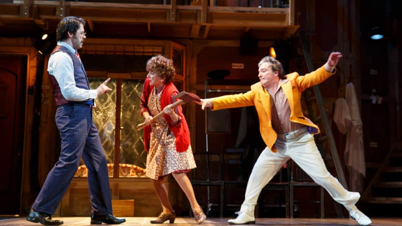 An older woman dressed in red tries to swing a fireman's axe towards a man in a business suit. A man in a yellow coat tries to pull her arm away.