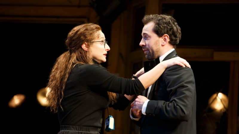 A stressed woman with glasses dressed in black puts her arms on the shoulders of a man dressed in a tux.