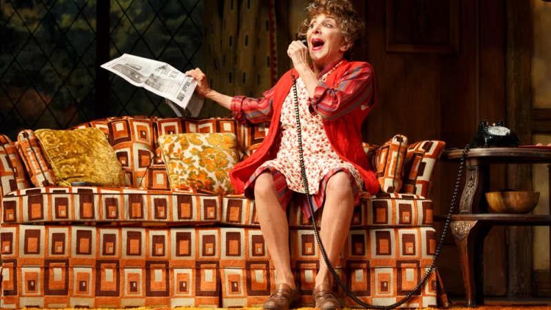 An older woman dressed in red sits on a brightly patterned couch. She excitedly speaks into a telephone and holds a newspaper in her other hand.