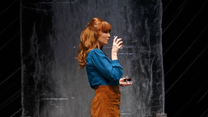 A woman in a blue blouse and copper skirt with long ginger hair stands in front of a mental background and drinks from a glass.