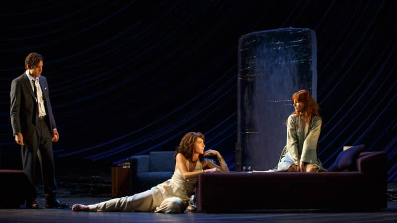 A woman sits on a bed and another woman on the floor leans onto the bed. A man off to the side watches them both. The room is dimly lit.