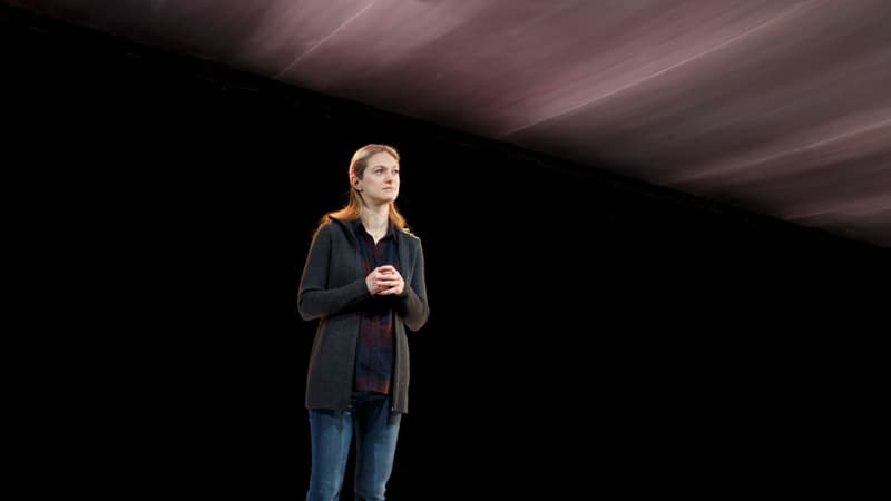 A blonde woman wearing a flannel under a zip up hoodie and jeans stands on stage, the background looking like a black abyss with no light.