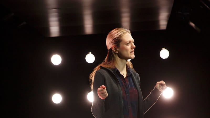 A blonde woman wearing a flannel under a zip up hoodie and jeans stands in front of a square panel with sixteen circular lights backlighting her.