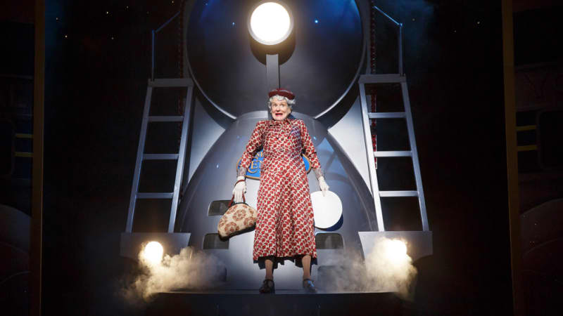 An older woman clutching a purse stands with her back up against a large, glowing train at night.