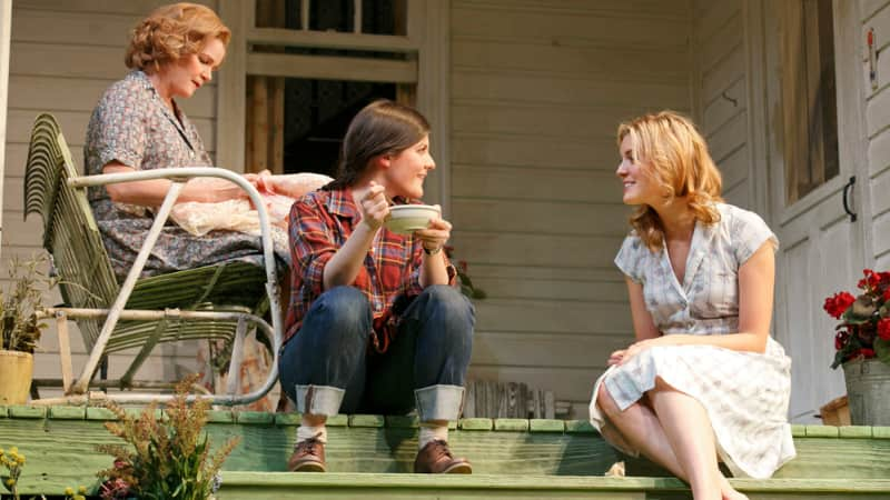 On an old green porch, a woman in a flannel and jeans eats from a bowl and chats with a barefoot woman in a dress. Behind them, another woman sits in a chair and mends a dress.